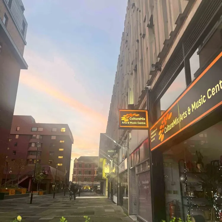 Urban street at dusk with illuminated storefront for arts and music centre among tall buildings.