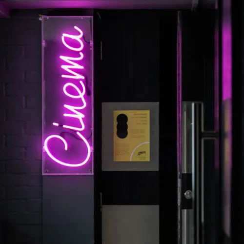 Magenta neon sign reading "Cinema" on dark wall beside framed poster and entrance door.