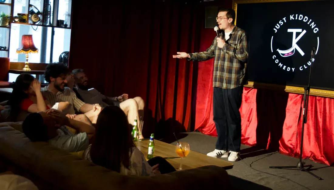 A comedian performs on stage with a microphone whilst seated audience members watch from the floor at an intimate comedy club venue.
