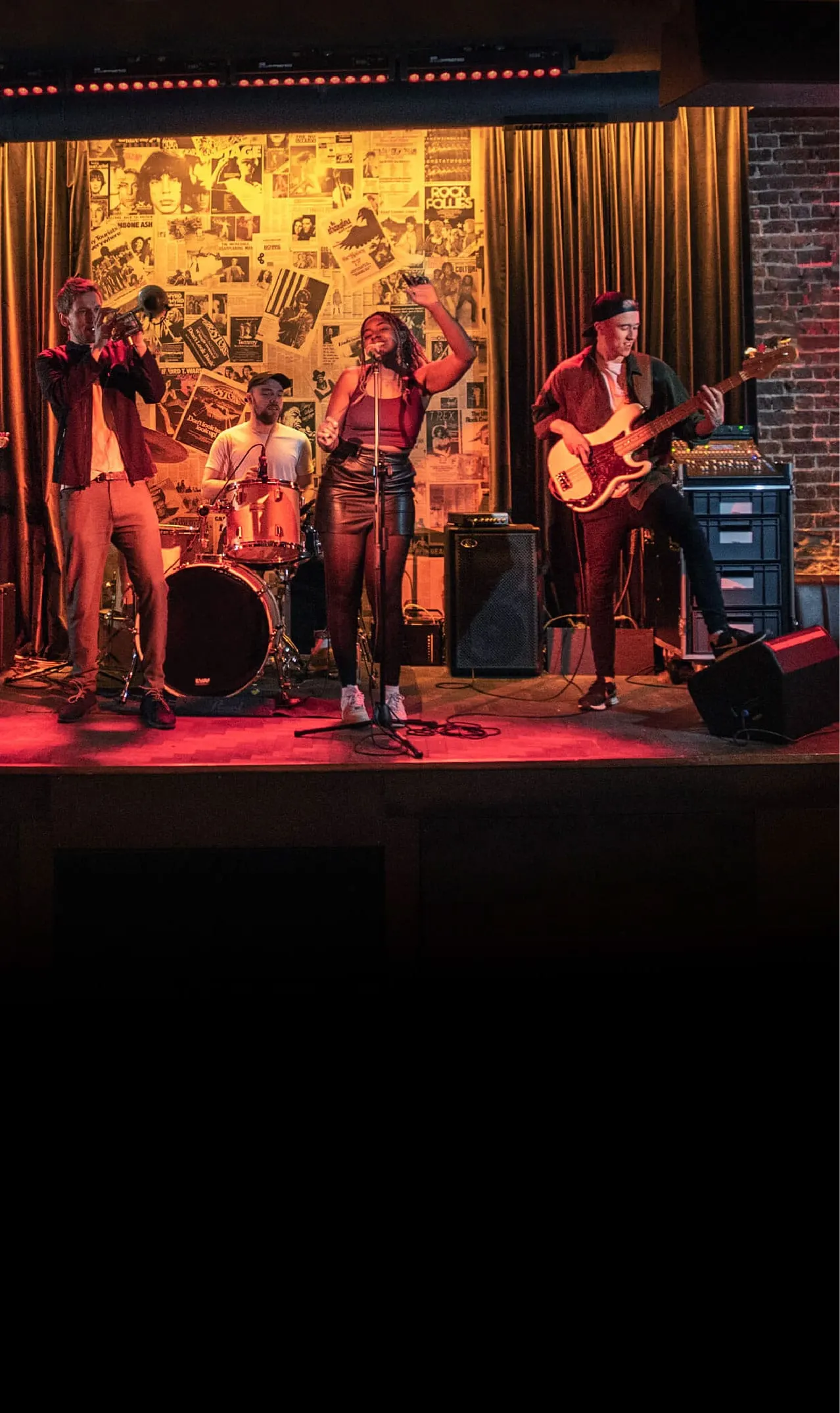 Four-piece band performing on stage with red lighting, featuring vocalist, drummer, bassist and guitarist against a backdrop of vintage music posters.