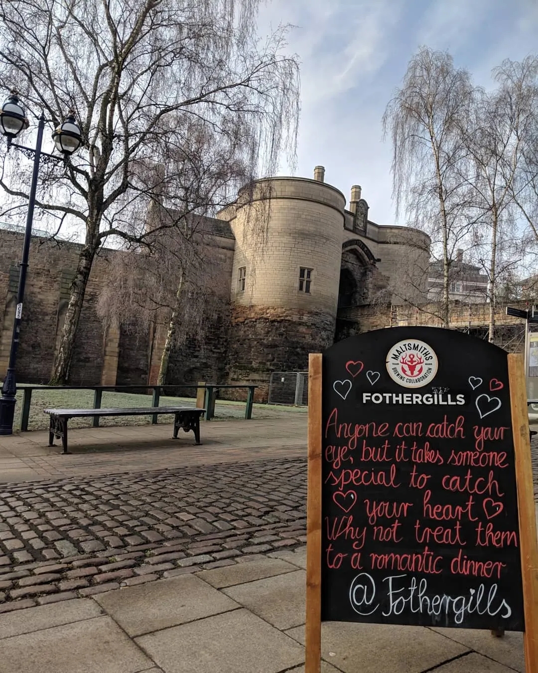 A romantic promotional sign stands in a paved courtyard with benches, bare birch trees and a historic stone fortress building behind it.