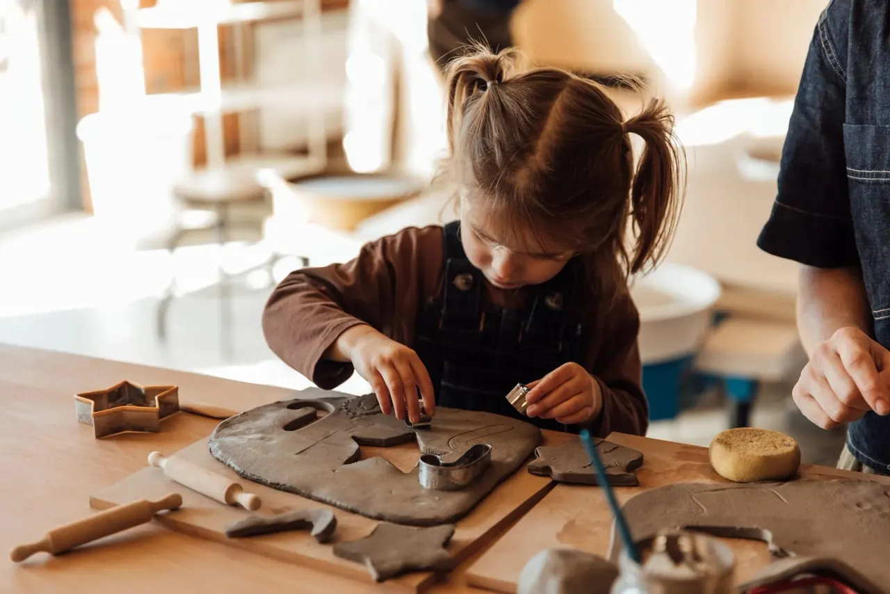 Young child doing a pottery or clay craft activity at a wooden table with an adult nearby.