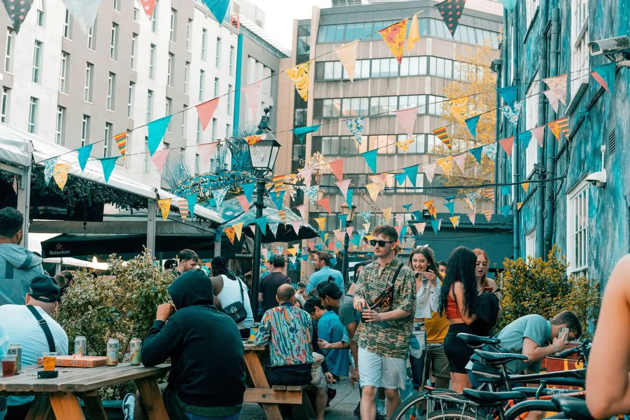 Urban courtyard festival with bunting and decorations, crowded with people of mixed ages socialising and mingling among buildings.