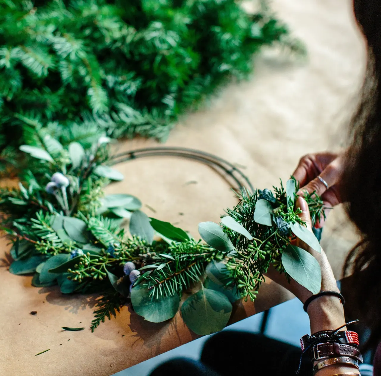 Person's hands arranging fresh evergreen foliage and eucalyptus leaves into a wreath on a sandy surface.