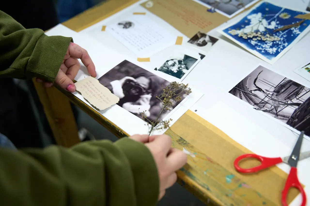 Person in green jumper arranging vintage photographs and handwritten notes on a wooden table during a creative workshop.