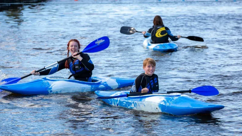 Children and young people kayaking in blue kayaks on calm water wearing wetsuits.