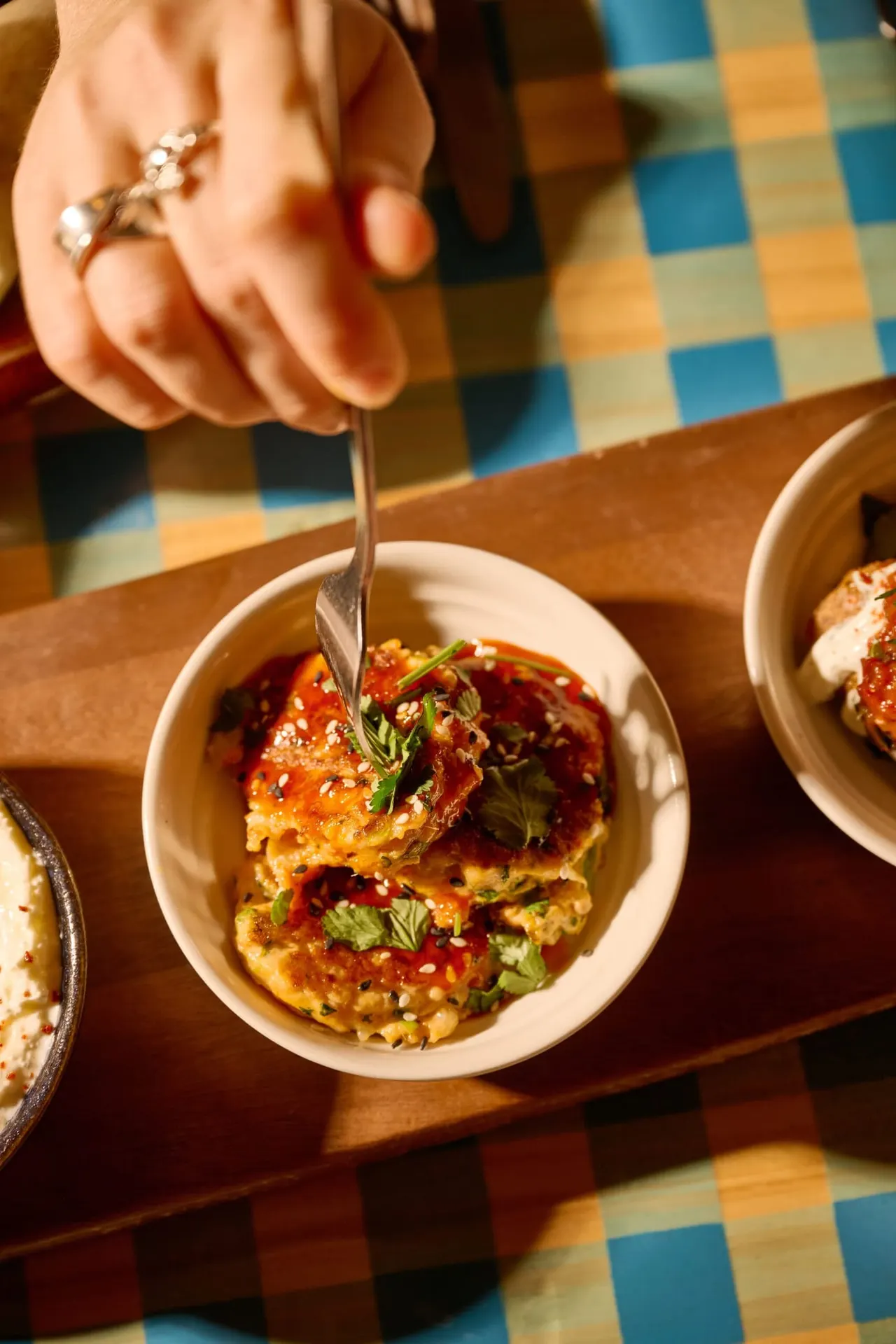 Person with jewelled hand holding spoon over bowl of glazed fritters with fresh herbs on checkered tablecloth.