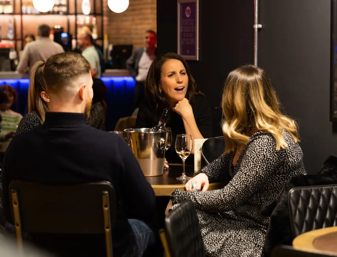 Three people socialising at a bar table with wine glasses and an ice bucket, in a modern venue with other guests in the background.