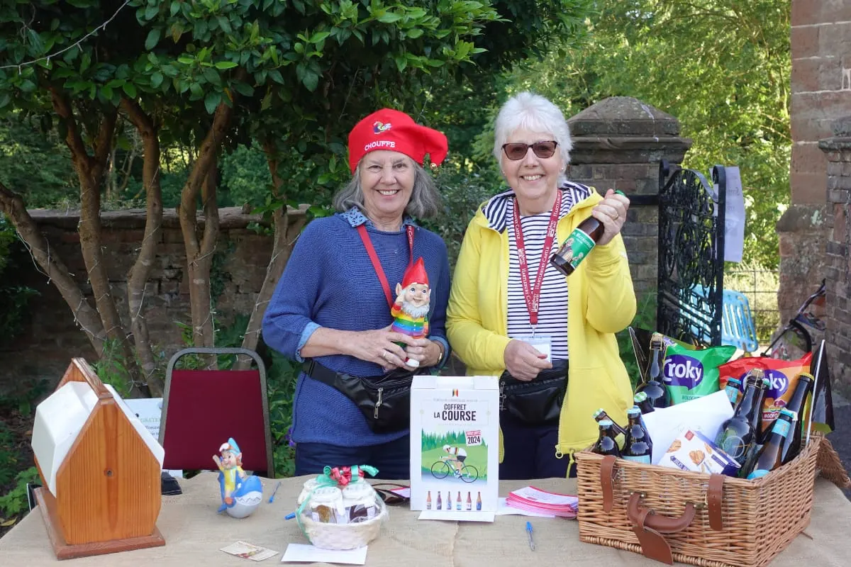 Two women at a garden stall wearing themed costumes and holding merchandise, with baskets and gnomes displayed on the table.