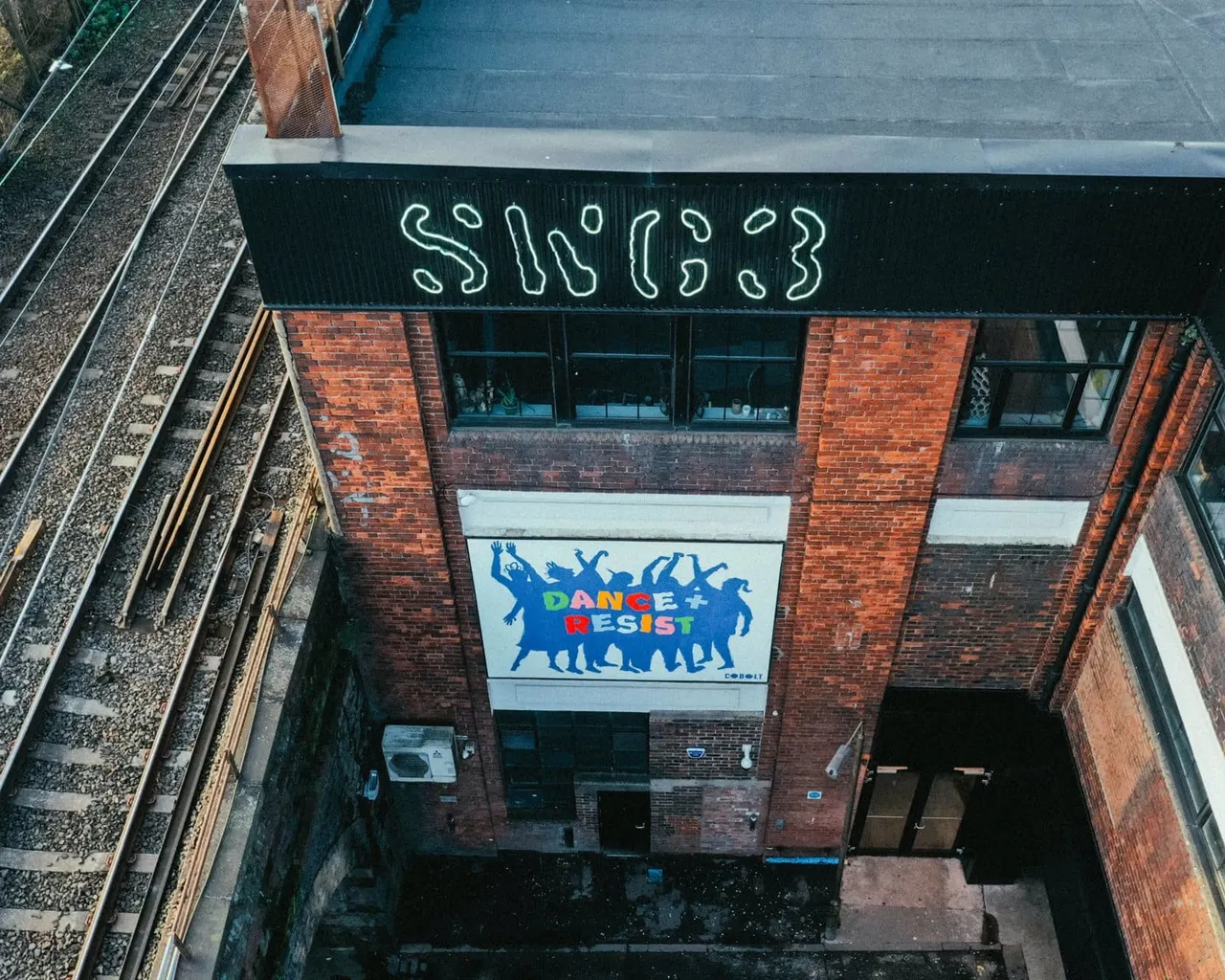 Overhead view of a brick building with neon signage reading "SWGJ3" and a "Dance + Resist" banner displayed on its façade.