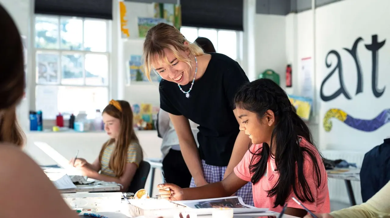 An instructor helps children with a painting or art activity at a bright, white-walled studio space with windows and artwork on the walls.
