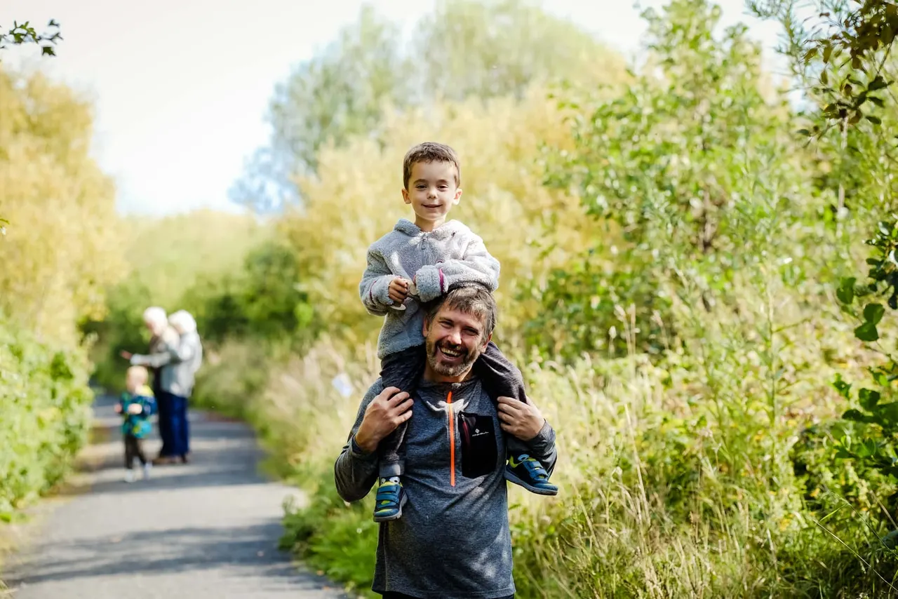 Man carrying young child on shoulders walking along tree-lined path with other visitors in background.