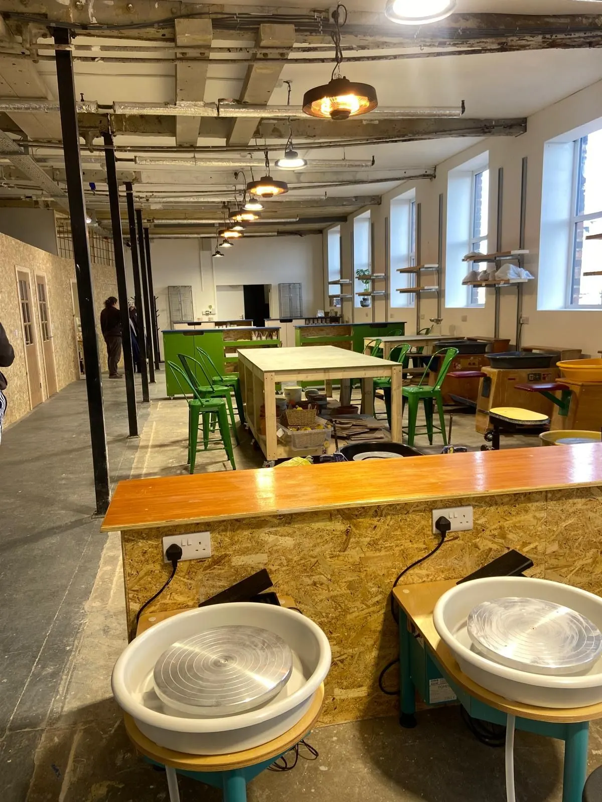 Industrial pottery studio with ceramic wheels in foreground and wooden tables with green chairs, large windows and exposed ceiling beams.