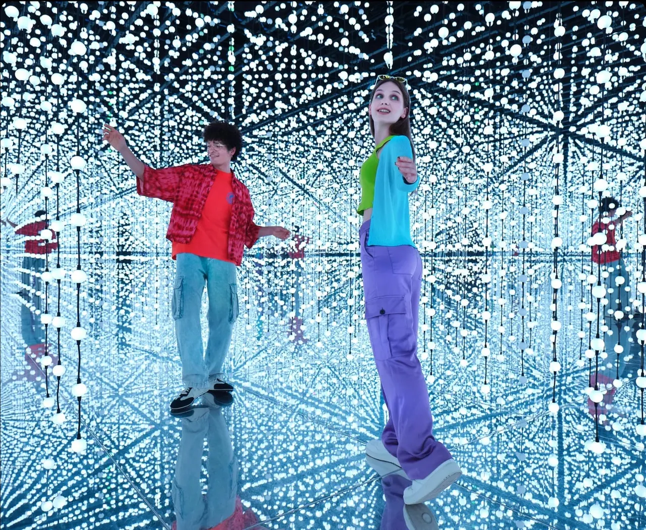 Two visitors explore an immersive digital room filled with thousands of glowing blue and white spheres.