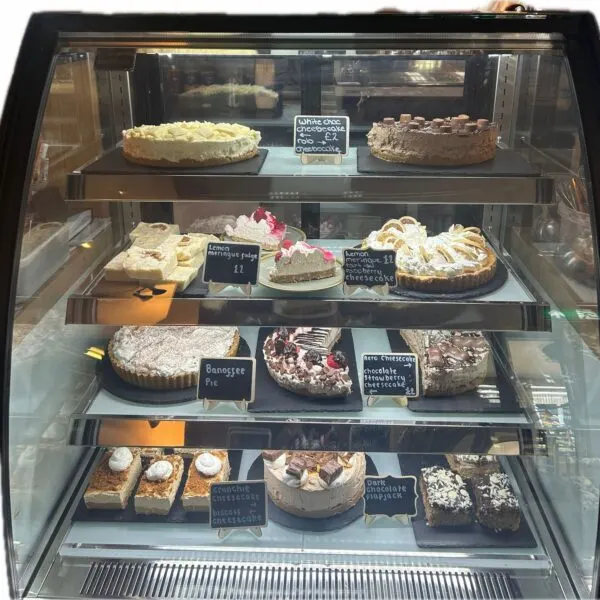 Refrigerated display case with multiple shelves of cakes and desserts with price labels.