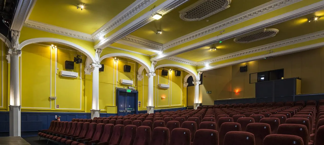 Cinema auditorium with rows of burgundy seats, ornate white columns and yellow walls with decorative cornicing.