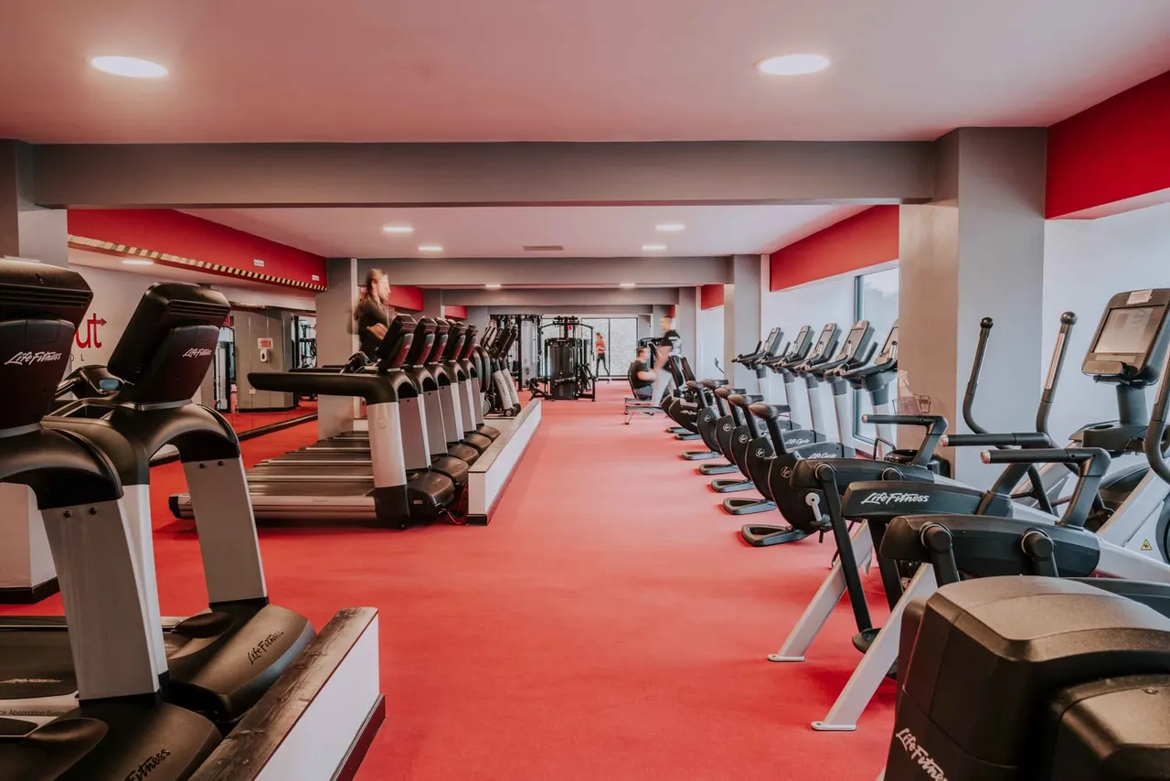 Spacious modern gym with red flooring and white walls featuring rows of cardio equipment and weight machines, with a few people exercising.