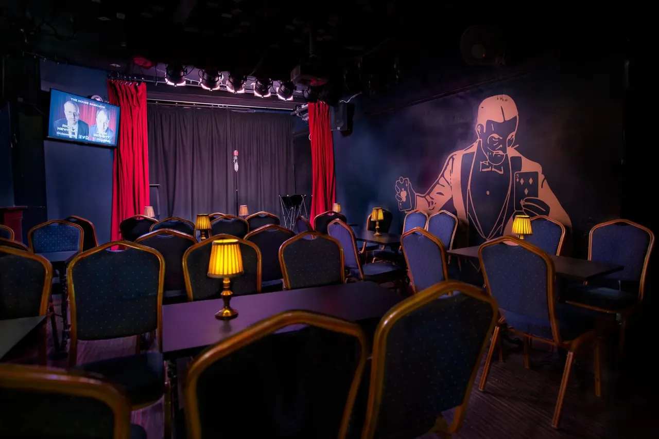 Cabaret-style theatre seating with tables and gold lamps facing a stage with red curtains and neon magician artwork.