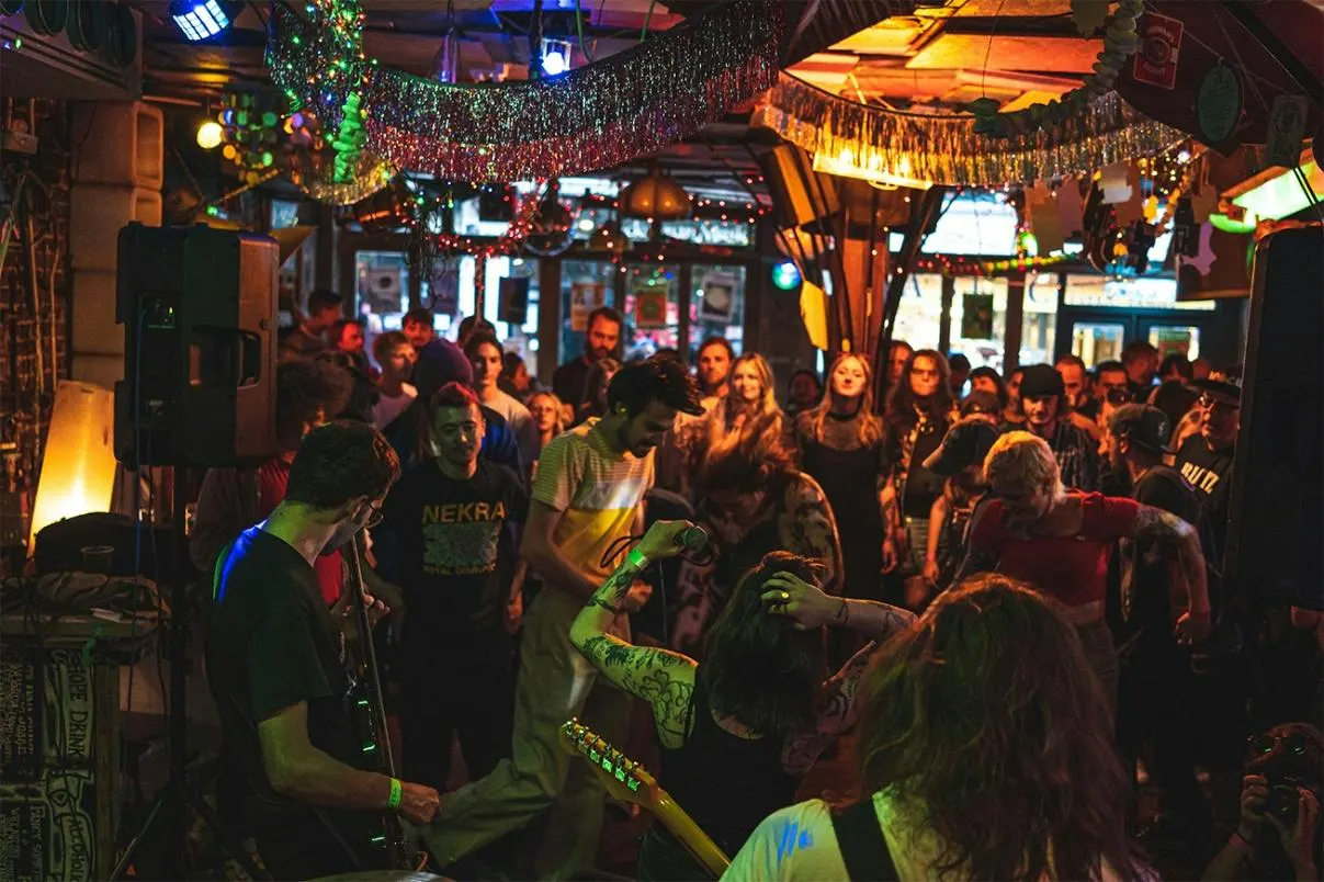 A crowded indoor venue with a live band performing to an audience under colourful overhead lights and tinsel decorations.