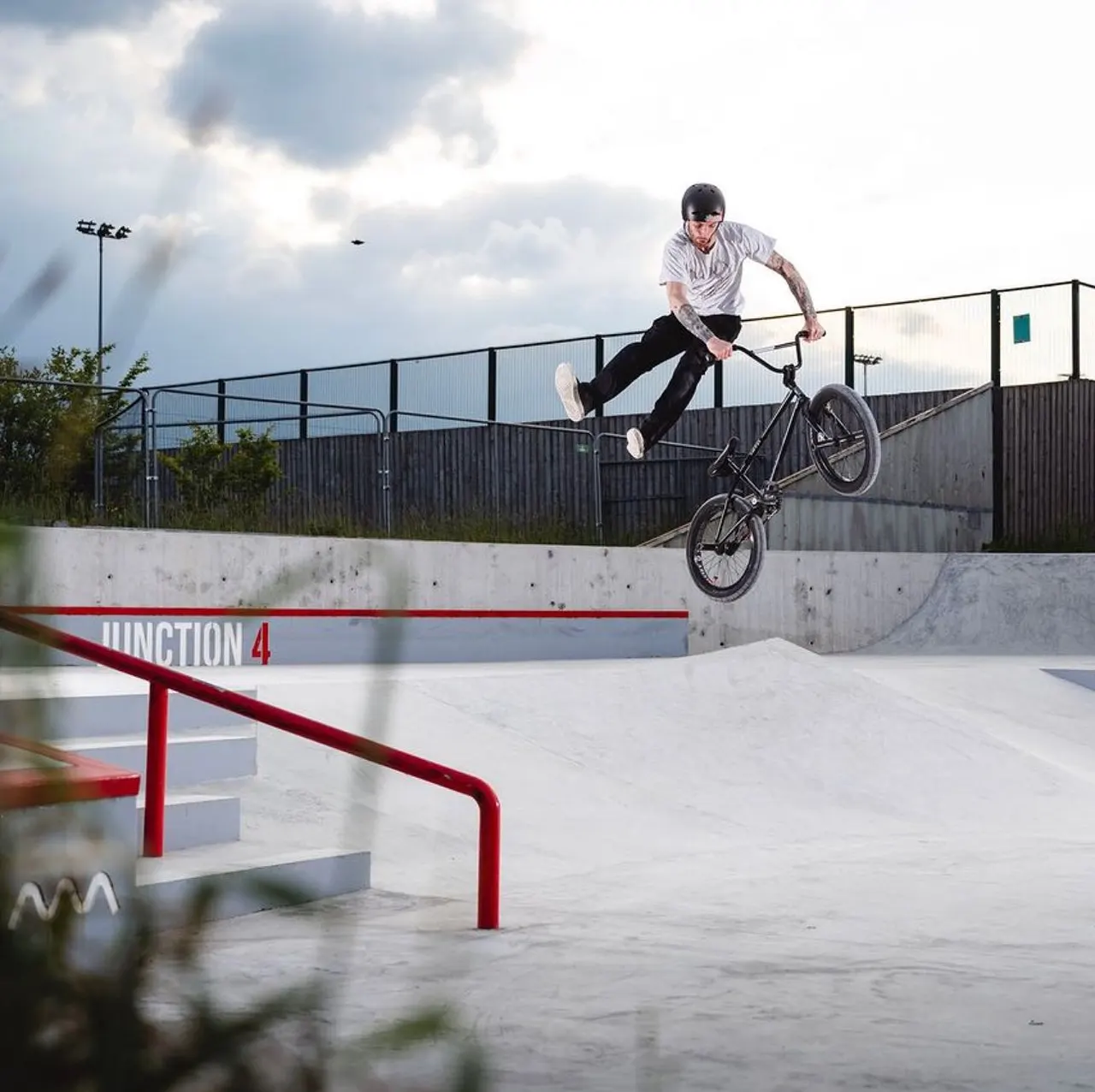 A young man performs a bike trick, jumping over a red rail at a concrete skate park.