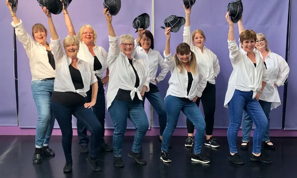 Group of ten people in white shirts and jeans holding up black hats against a purple backdrop on stage.