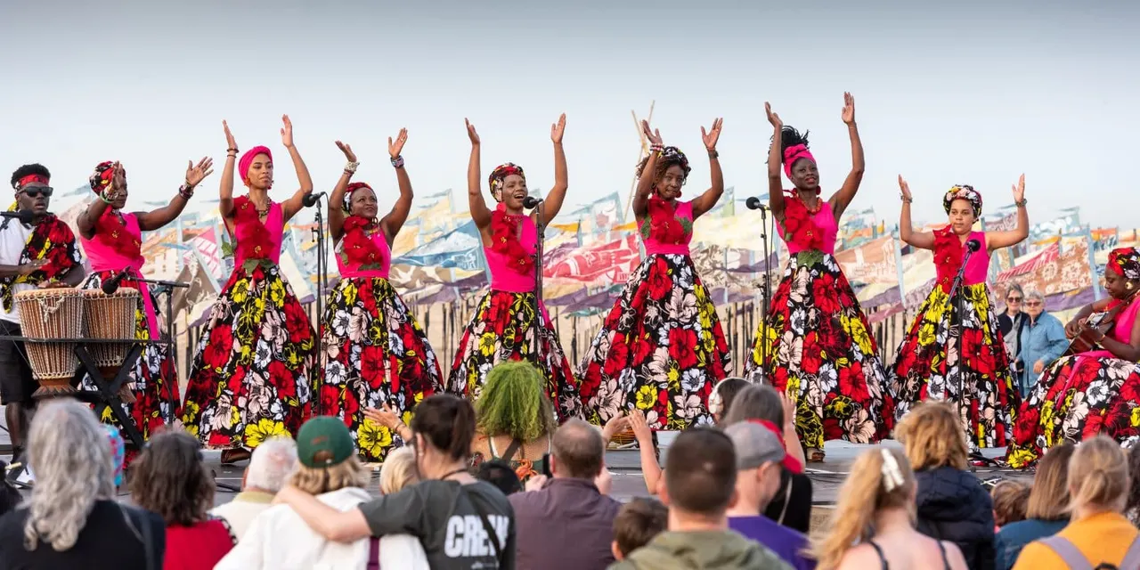Performers in bright pink and floral outfits dance with raised arms on stage whilst a crowd watches from below.