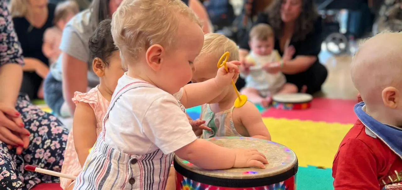 Toddlers and babies with adults participate in a music and movement session, playing a colourful drum together.