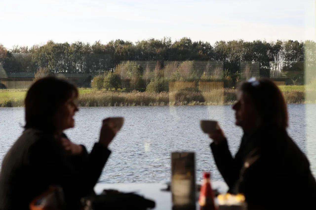Two people chatting by a waterside viewing area overlooking a lake, reeds and wooded landscape.