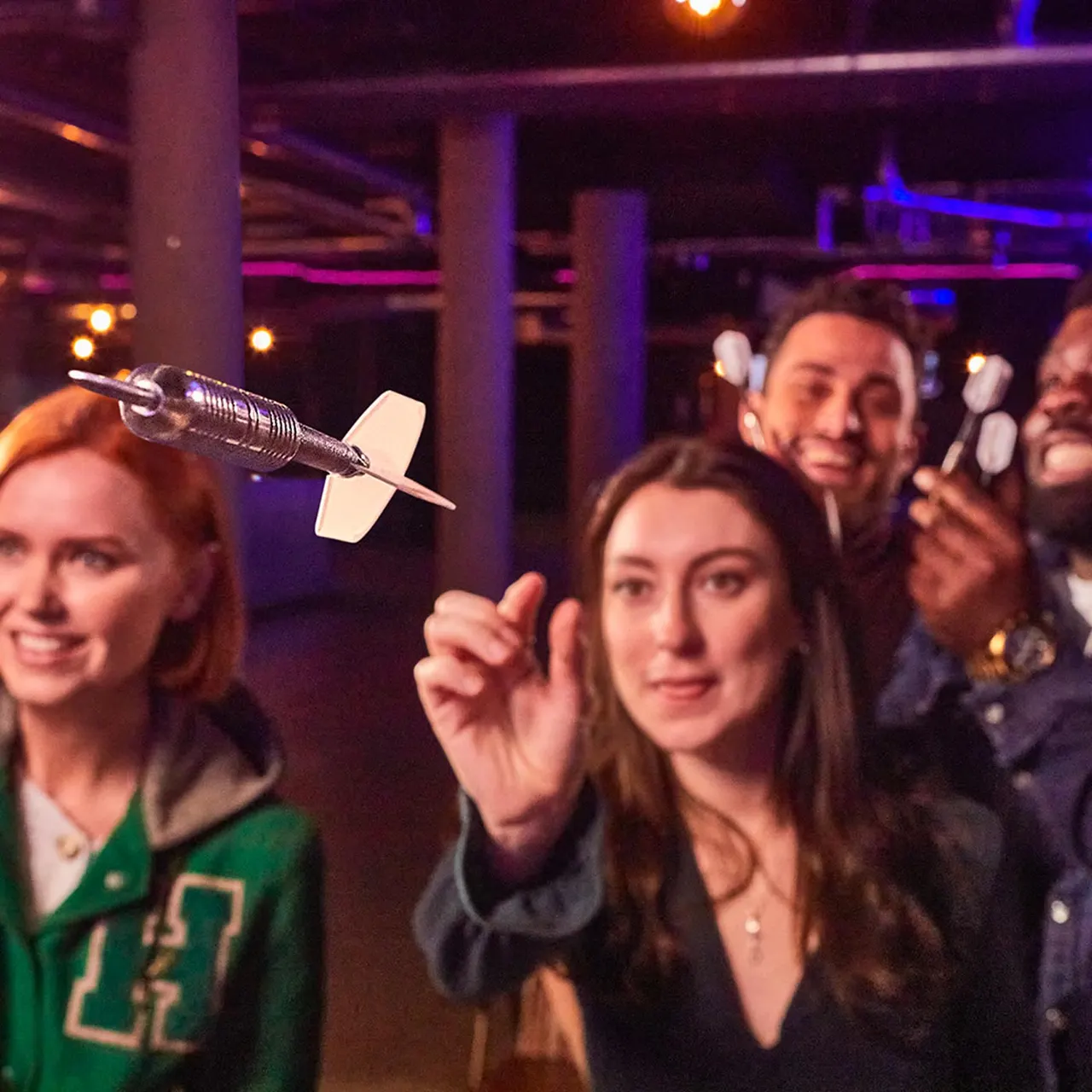 A woman holds up a dart at an indoor entertainment venue with coloured lighting, surrounded by smiling guests enjoying the atmosphere.