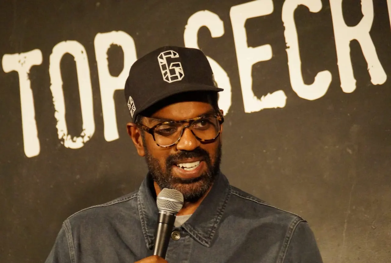 A man wearing a black cap and glasses holds a microphone whilst performing on stage in front of a dark backdrop with large white text.