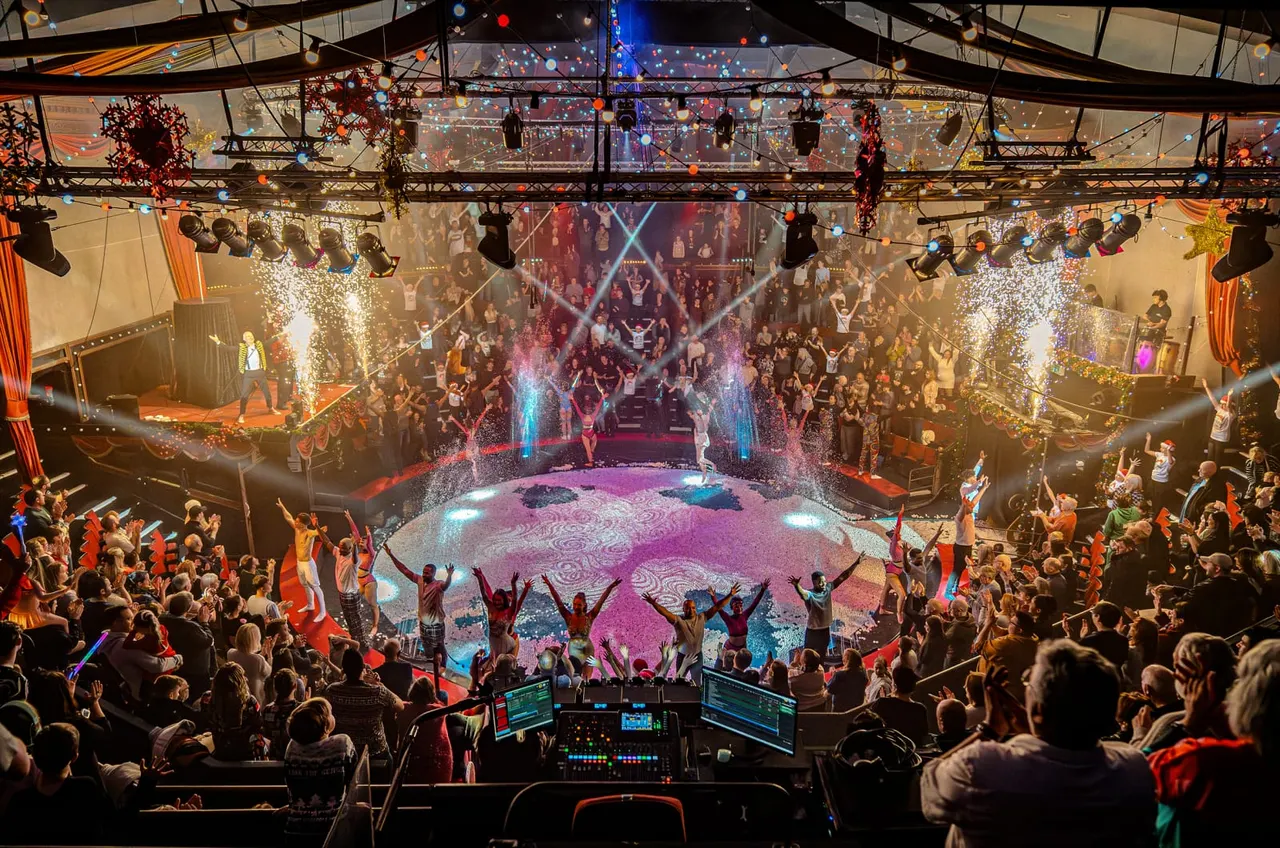 Aerial view of a packed circus tent with performers on a lit stage, audiences dancing and cheering, pyrotechnics, and colorful lighting throughout.