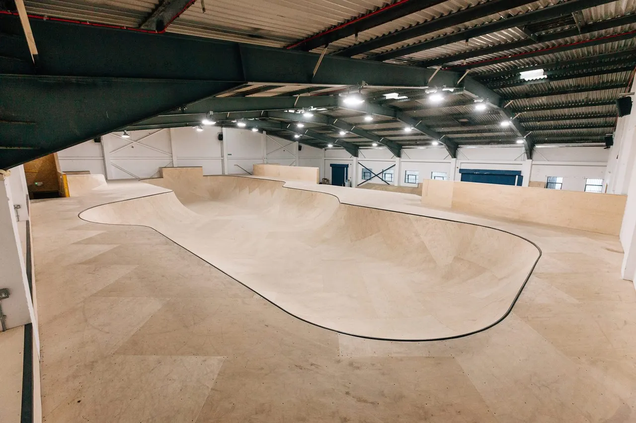 Indoor skate bowl and ramps in a spacious warehouse with concrete floor and industrial lighting.