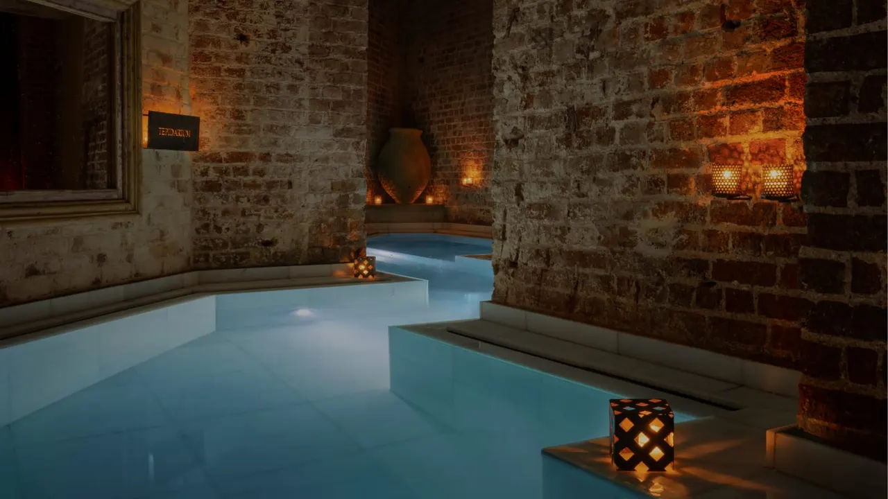 Interior of spa with turquoise thermal pools set within exposed brick stone walls, lit by warm ambient lighting and decorative lanterns.