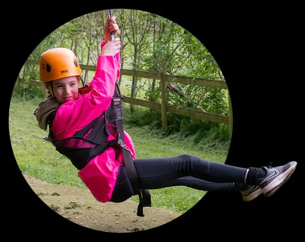 Child in orange helmet and pink jacket zip-lining through woodland on an adventure course.