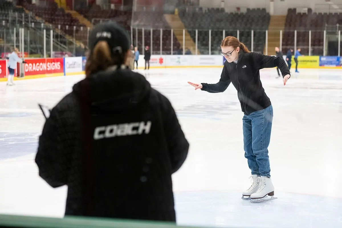 A coach in a black jacket watches a young woman ice skating on an indoor rink.