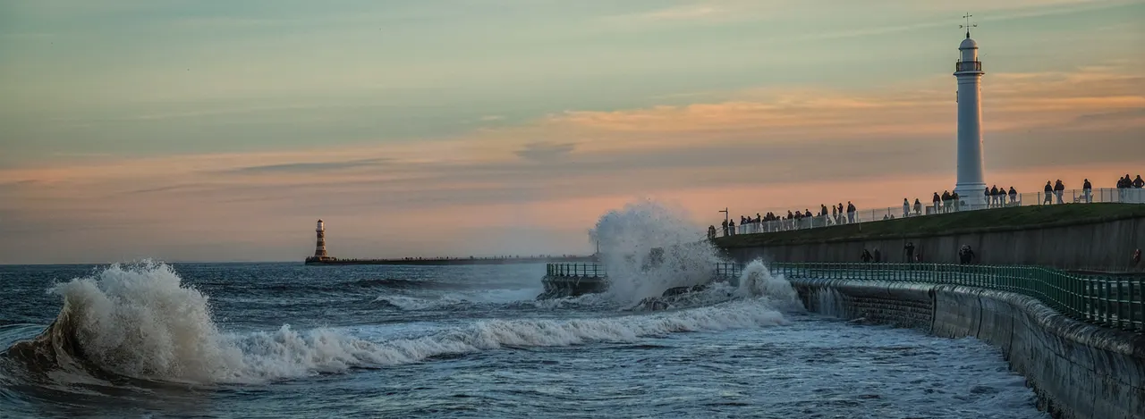 Large waves crash against a seafront pier with a lighthouse and spectators during sunset.