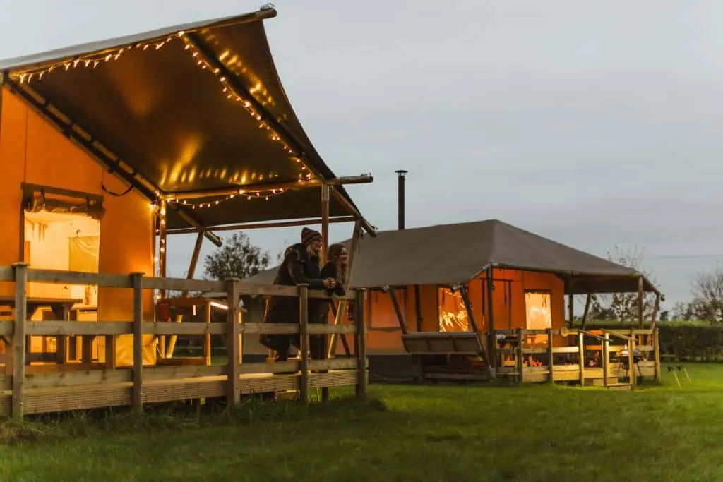 Illuminated glamping tents with canopies and decking at dusk, two people sitting on the veranda of the nearest tent.