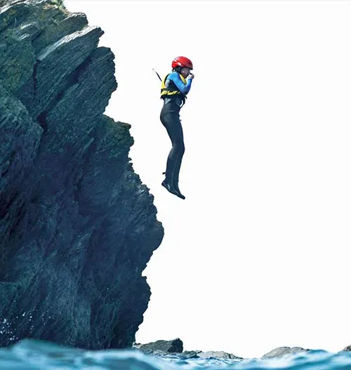 Person cliff jumping in red helmet and blue jacket above turquoise water near rocky cliff.