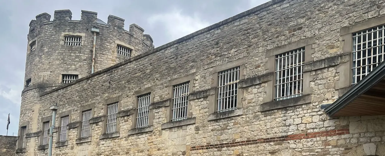 Historic stone castle building with crenellated tower and barred windows under cloudy sky.