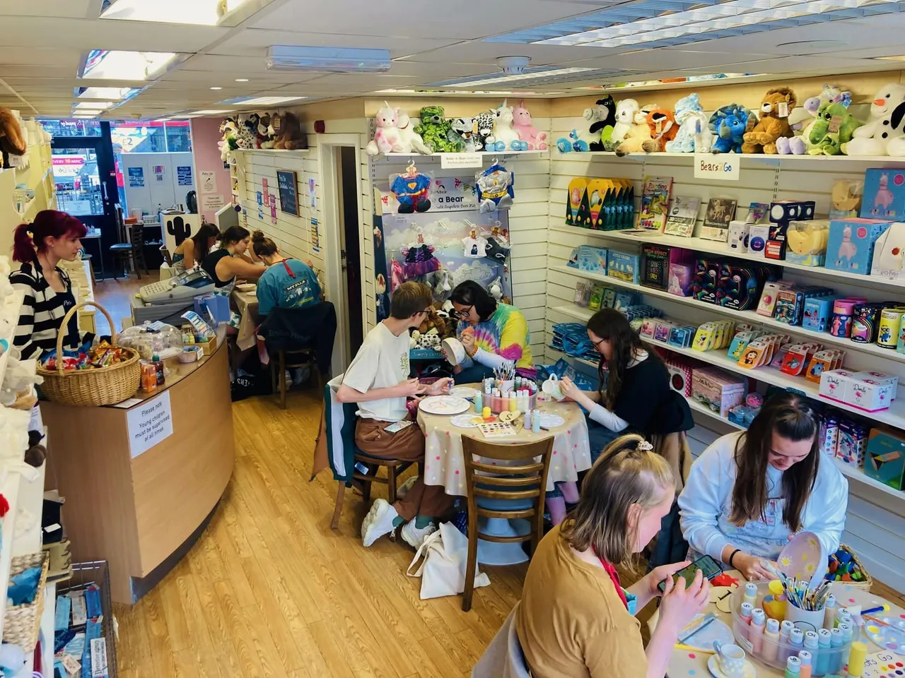 Children and adults painting pottery at tables in a bright ceramic studio with shelves of stuffed animals and craft supplies.