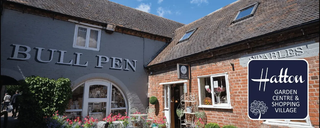 Historic stone and brick buildings at a countryside leisure venue, with flowering planters and signage visible on a sunny day.