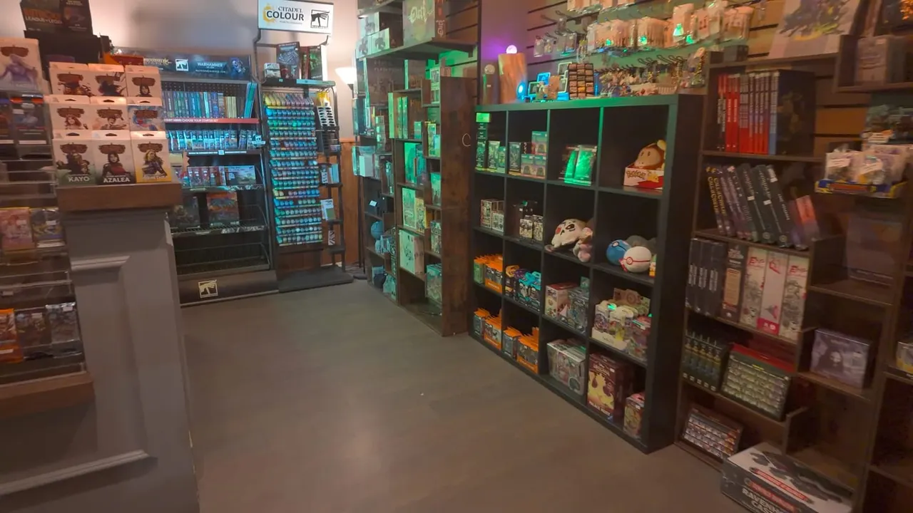 Interior of a gaming and collectibles shop with dark shelving units displaying merchandise, illuminated with green neon lighting.