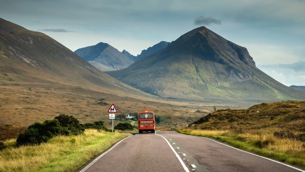 Red tour bus driving on a Highland road towards dramatic mountains in a remote Scottish landscape.