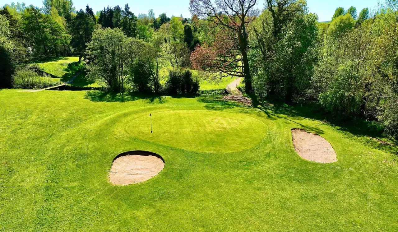 Golf green with bunkers and a flag, surrounded by trees and rolling grassland.