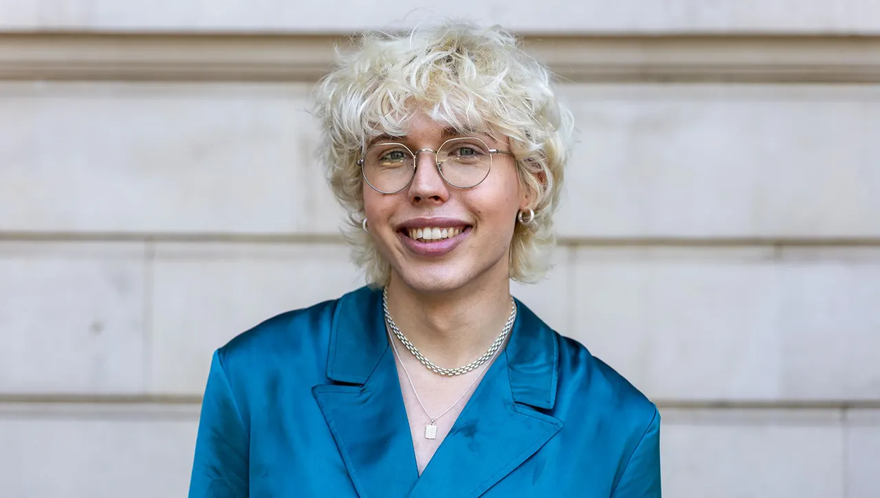This is a portrait photograph of a smiling person with short white-blonde curly hair, wearing glasses, a blue jacket and chain necklace against a white panelled background.