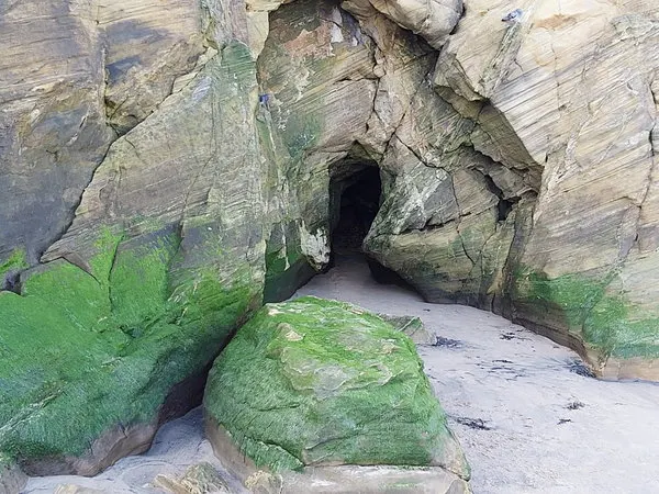 Rock cave entrance with sand floor and moss-covered boulders on a beach.
