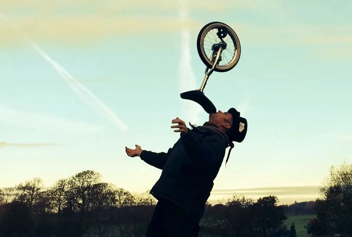 Person performing a BMX bike trick in mid-air against a pale sky with rural landscape below.