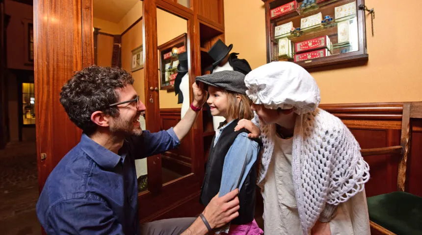 A man adjusts a child's hat whilst a woman watches in a museum gallery with wooden panelling and period clothing displays.