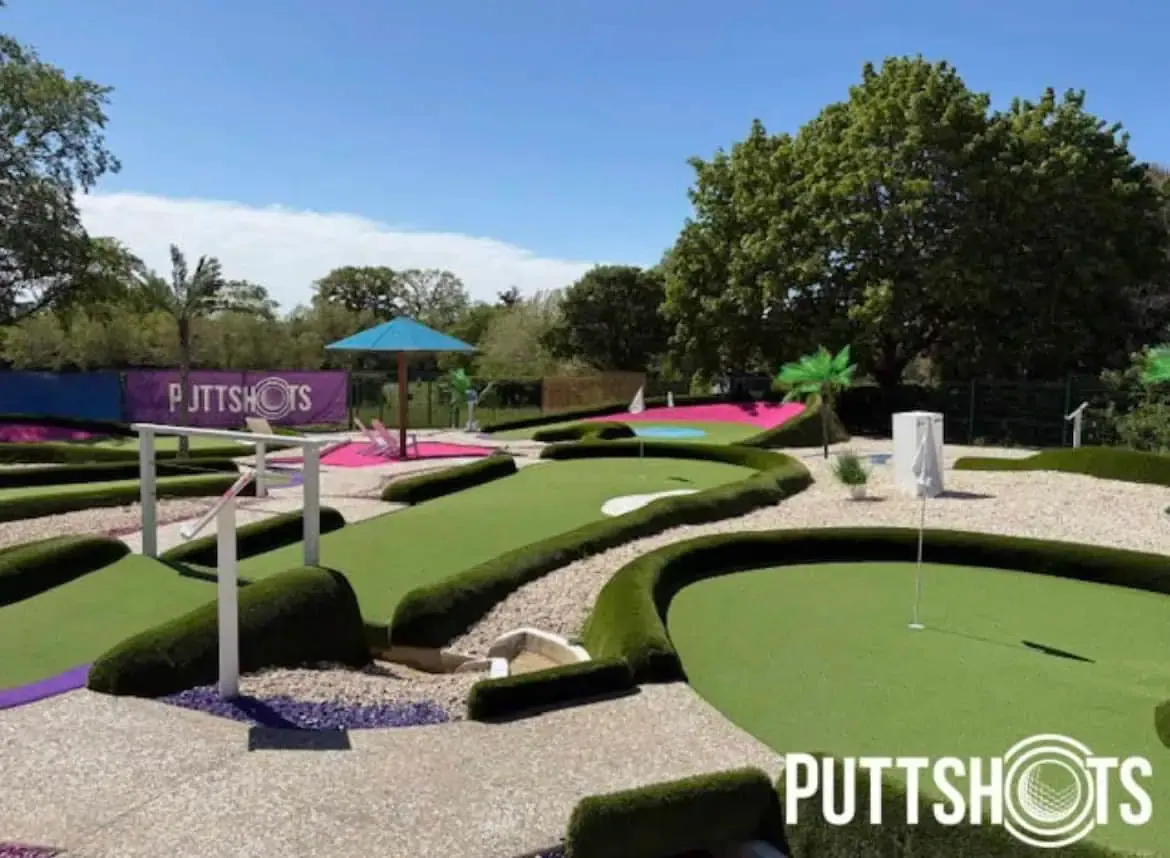 Colourful mini golf course with green artificial putting surfaces, hedge features and a blue umbrella, surrounded by mature trees under clear sky.