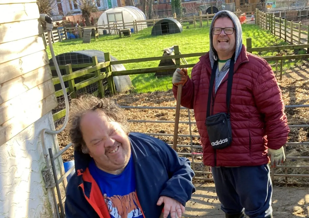 Two visitors smiling at a city farm with fenced enclosures, polytunnels and farm buildings in the background.